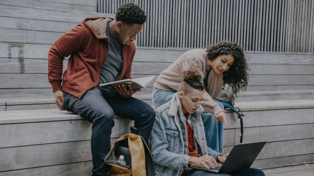 Three university students sitting on campus stairs doing group study, showing how to stay motivated for study through teamwork.